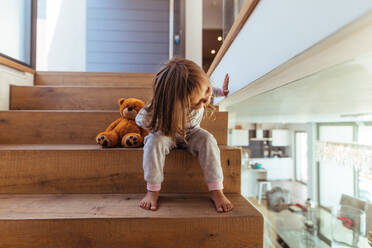 Nettes kleines Mädchen sitzt auf einer Treppe und schaut durch eine Glaswand nach unten. Mädchen Kind späht in Wohnzimmer von Holztreppe zu Hause. - JLPSF21443