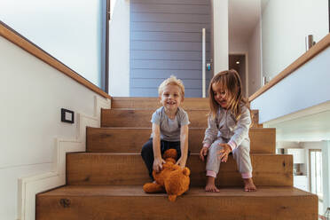 Kleine Kinder sitzen auf einer Treppe, ein Junge hält einen Teddybär in der Hand. Geschwister spielen mit einem Teddybären auf einer Treppe zu Hause. - JLPSF21446