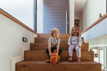 Fröhliche Kinder sitzen auf einer Treppe mit einem Teddybär. Lachender kleiner Junge und Mädchen sitzen auf einer Holztreppe mit Teddybär. - JLPSF21447