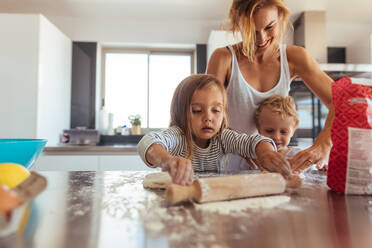 Mutter und kleine Kinder backen Kekse in der Küche. Geschwister mit Nudelholz und Mehl helfen der Mutter in der Küche zu Hause - JLPSF21450