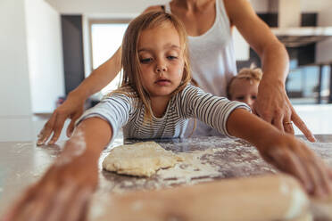 Beautiful young girl learning cooking and baking with flour and a rolling pin in a kitchen at home with her mother and brother standing by. - JLPSF21451