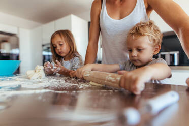 Kleiner Junge mit seiner Mutter und Schwester bereitet Teig mit Nudelholz auf dem Küchentisch vor. Mutter mit Kindern beim Kekse backen in der Küche. - JLPSF21452