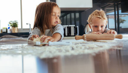 Ein kleiner Junge benutzt ein Nudelholz und ein Mädchen knetet den Teig mit Mehl auf dem Küchentisch. - JLPSF21453