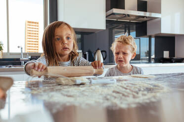 Niedliches kleines Mädchen, das einen Teig für Kekse zubereitet und ein weinender kleiner Junge, der daneben steht. Kinder machen Kekse in der Küche. - JLPSF21454