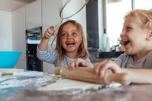 Lächelndes kleines Mädchen, das ein Sieb mit einem Jungen hält, der Teig mit einem Nudelholz auf dem Küchentisch herstellt. Kinder haben Spaß beim Kekse backen in der Küche. - JLPSF21457