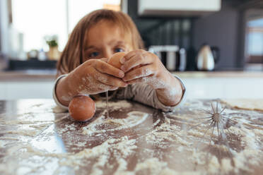 Kleines Mädchen bricht ein Ei auf dem Küchentisch mit Mehl bedeckt. Mädchen Kind lernen Kochen in der Küche zu Hause und machen ein Chaos. - JLPSF21460