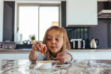 Niedliches kleines Mädchen schlägt ein Ei in eine kleine Pfanne auf dem Küchentisch auf. Schönes junges Mädchen backt in der Küche. - JLPSF21461