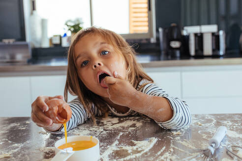 Unschuldiges junges Mädchen, das ein Ei in eine kleine Pfanne schlägt und den Teig probiert. Niedliches junges Mädchen beim Backen in der Küche. - JLPSF21462