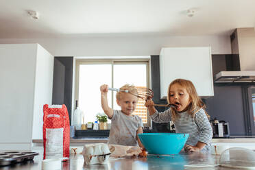 Young girl licking batter from spatula with her brother holding a whisk. Siblings making cake batter in kitchen. - JLPSF21466