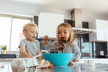 Kids preparing chocolate cake batter with girl licking batter from spatula with her brother holding a whisk. Small kids preparing cake batter in kitchen. - JLPSF21469