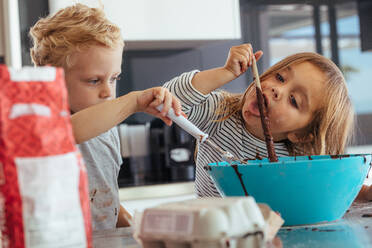 Little kids mixing batter in a bowl for baking, with girl licking a spoon. Children baking in the kitchen. - JLPSF21470