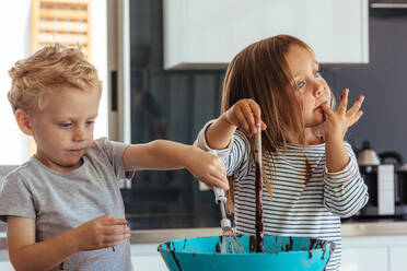 Little boy and girl mixing batter in a bowl for baking, with girl licking her fingers. Small kids baking in the kitchen. - JLPSF21471