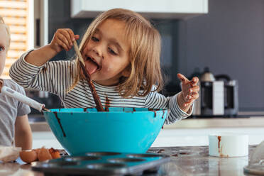 Little girl licking spoon while mixing batter for baking in kitchen and her brother standing by. Cute little children making batter for baking. - JLPSF21472