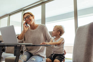 Kleiner Junge sitzt mit seiner Mutter und spricht mit dem Handy. Beschäftigte Frau mit Sohn im Büro. - JLPSF21492