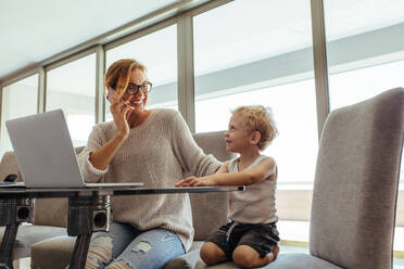 Young mother with son working from home, talking on the phone and smiling. Busy young woman with son working in home office. - JLPSF21493