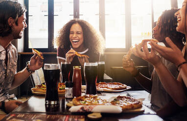 Young woman eating pizza and laughing while sitting with her friends in a restaurant. Group of friends enjoying while having food and drinks at cafe. - JLPSF21496