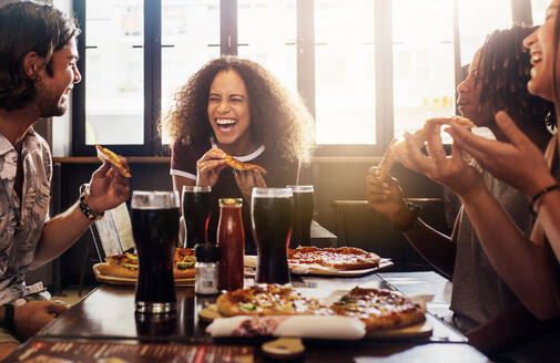 Junge Frau isst Pizza und lacht, während sie mit ihren Freunden in einem Restaurant sitzt. Eine Gruppe von Freunden genießt das Essen und die Getränke in einem Café. - JLPSF21496
