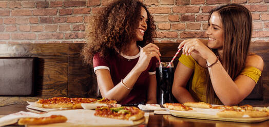 Zwei Freundinnen in einem Restaurant schauen sich an und teilen sich ein kaltes Getränk mit zwei Strohhalmen. Fröhliche Freundinnen beim gemeinsamen Mittagessen im Restaurant. Frauen mit Pizza und Cola auf dem Tisch in einem Schnellrestaurant. - JLPSF21503