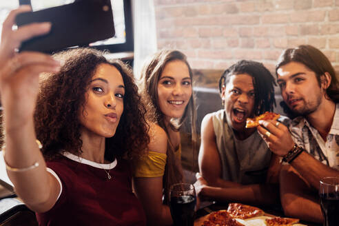 Eine Frau macht ein Selfie mit Freunden beim Pizzaessen in einem Restaurant. Eine Gruppe von Männern und Frauen genießt das Essen und macht ein Selfie im Restaurant. - JLPSF21511