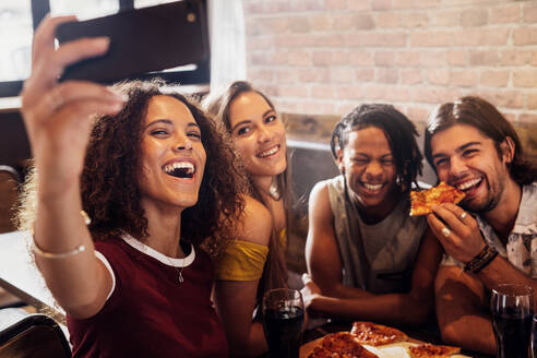 Gruppe glücklicher multirassischer Freunde, die ein Selfie mit einem Smartphone machen, während sie eine Party in einem Pizzarestaurant feiern. Aufgeregte Freunde, die ein Selfie mit einem Telefon machen, während sie im Restaurant abhängen. - JLPSF21512