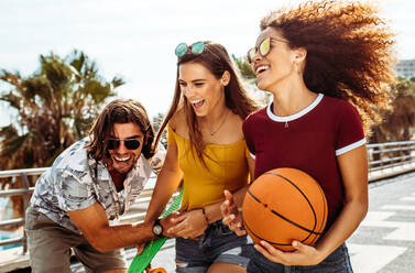 Gruppe von drei jungen Freunden, die sich im Freien vergnügen. Frauen mit Basketball und Skateboard lachen mit einem männlichen Freund. - JLPSF21515