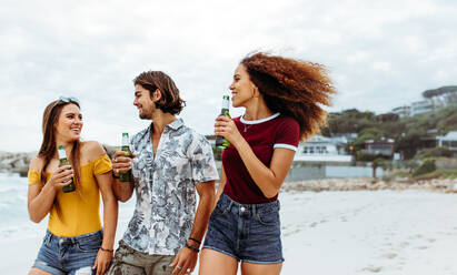 Gruppe von drei jungen Freunden, die mit Bierflaschen im Freien spazieren gehen. Multi-ethnische Freunde mit Bier, die am Strand spazieren gehen. - JLPSF21545