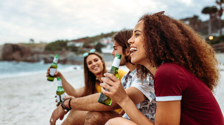 Lächelnde junge Frau mit Bierflasche und Freunden, die am Strand sitzen. Seitenansicht einer Gruppe von Freunden, die am Abend eine Party am Strand feiern. - JLPSF21546