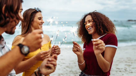 Eine Gruppe junger Freunde feiert den Neujahrstag am Strand. Eine Gruppe von Männern und Frauen hat Spaß mit Wunderkerzen im Freien am Strand. - JLPSF21550