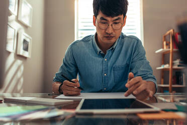 Kreativer Mann arbeitet im Büro mit Tablet-PC und dawning Buch. Männliche Innenarchitekt macht eine Zeichnung auf Buch, während die Referenz von digitalen Tablet an seinem Schreibtisch. - JLPSF21563