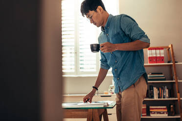 Mann mit Kaffee am Tisch stehend, der einen Bauplan betrachtet, Architekt arbeitet an seinem Schreibtisch. - JLPSF21567