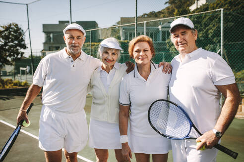 Ältere Männer, die Tennisschläger und Ball halten, stehen mit ihren Partnerinnen zusammen. Senioren stehen zusammen und halten sich gegenseitig, um gemischtes Doppel zu spielen. - JLPSF21572
