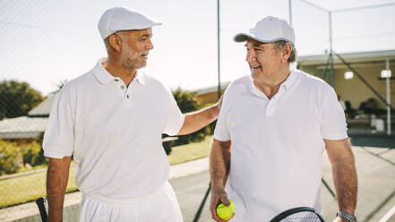 Ältere Männer, die während eines Tennisspiels auf einem Tennisplatz spazieren gehen. Lächelnde ältere Männer in Tenniskleidung, die sich miteinander unterhalten und Tennisschläger und Ball halten. - JLPSF21577