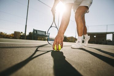 Ground level view of a man picking a tennis ball on a sunny day. Low angle view of a tennis player bending forward to pick the ball with sun in the background. - JLPSF21581