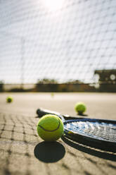 Ground level shot of a tennis racket with balls on tennis court. Tennis racket and balls placed beside a net on a tennis court on a sunny day. - JLPSF21584