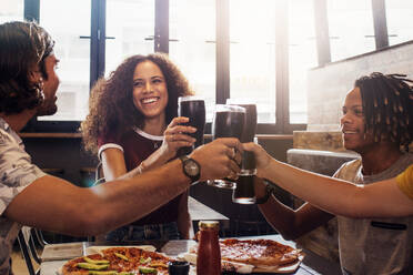 Lächelnde junge Männer und Frauen sitzen in einem Restaurant und stoßen mit alkoholfreien Getränken an. Multi-ethnische Gruppe von Menschen stößt mit kalten Getränken und Pizza auf einem Tisch im Restaurant an. - JLPSF21595