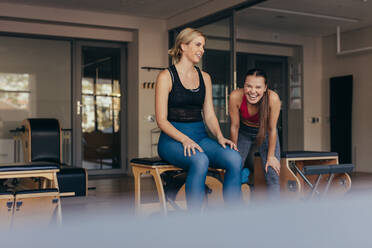 Frauen sitzen auf einer Bank im Fitnessstudio und entspannen sich, während ihr Trainer lacht. Frauen machen Pilates-Training im Fitnessstudio. - JLPSF21619