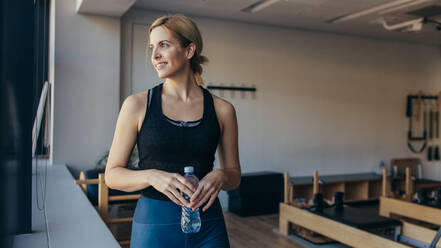 Lächelnde Fitness-Frau, die eine Wasserflasche in einem Pilates-Fitnessstudio hält. Frau entspannt sich nach dem Training in einem Pilates-Fitnessstudio. - JLPSF21645