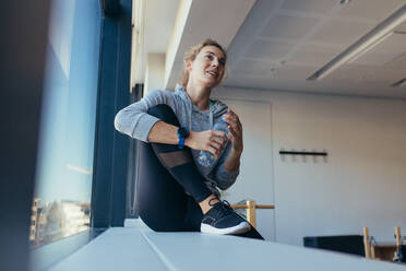 Frau entspannt sich nach dem Training am Fenster in einem Pilates-Fitnessstudio. Fitness-Frau hält eine Wasserflasche in einem Pilates-Fitnessstudio. - JLPSF21646