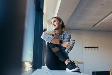 Smiling fitness woman holding water bottle sitting in a pilates gym. Woman relaxing after workout looking out of a window in a pilates gym. - JLPSF21647