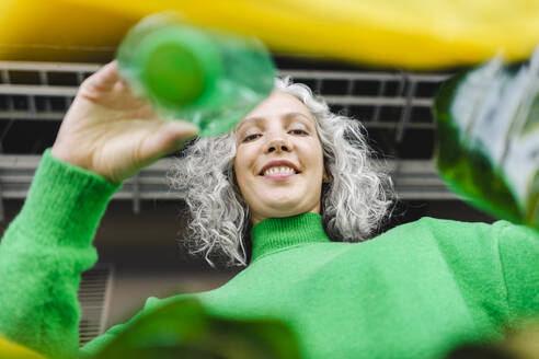 Lächelnde Frau mit grauem Haar, die eine grüne Flasche in eine gelbe Plastiktüte steckt - SEAF01479