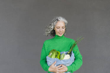 Frau mit grauem Haar betrachtet Netzbeutel mit frischem Gemüse vor einer Wand - SEAF01486