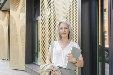 Smiling businesswoman with laptop standing in front of wall - SEAF01495