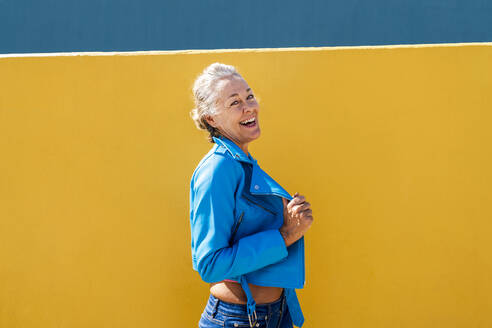 Happy woman wearing blue leather jacket by colored wall - OIPF02601