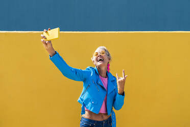 Happy mature woman gesturing and taking selfie through mobile phone in front of colored wall - OIPF02617