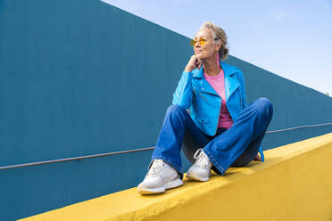 Reife Frau in blauer Lederjacke sitzt auf gelber Wand - OIPF02625