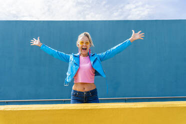 Happy mature woman with arms outstretched in front of blue wall - OIPF02630