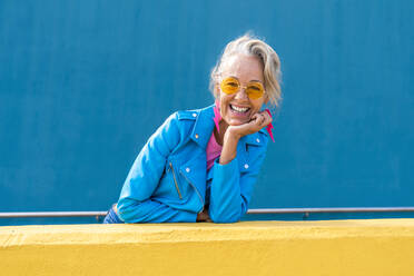 Glückliche Frau mit Sonnenbrille vor einer blauen Wand - OIPF02635