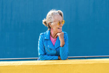 Glückliche reife Frau mit Hand am Kinn vor einer blauen Wand - OIPF02636