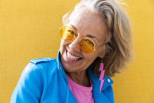 Glückliche reife Frau mit Sonnenbrille vor einer gelben Wand - OIPF02640