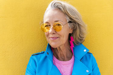 Smiling mature woman wearing sunglasses leaning on yellow wall - OIPF02644